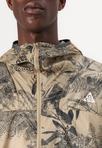 Beige Jacke mit schwarzen Blattmustern, verstellbarer Kapuze und Reißverschluss. Mit einem kleinen weißen Logo auf der linken Brustseite. Glatte Textur.