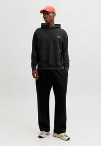 Must kapuutsiga dressipluus esitaskuga, kombineeritud lahtiste mustade pükstega ja beežide tossudega. Modellil on peas korallikarva pesapallimüts.