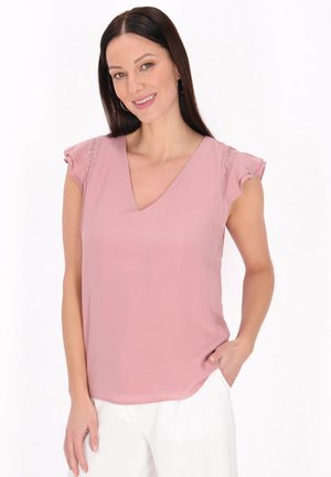 Donna con capelli lunghi e scuri che indossa una camicetta rosa senza maniche con dettagli in pizzo e pantaloni bianchi, sorridente con una mano in tasca.