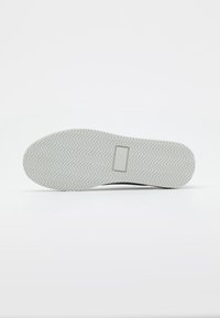 Semelle de chaussure avec un motif de zigzag gris clair et un petit patch logo rectangulaire au centre sur fond blanc.