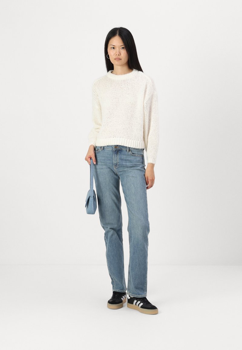 Pull en tricot crème avec un col rond, associé à un jean droit bleu clair et des baskets noires avec des rayures blanches. Sac à main bleu clair.