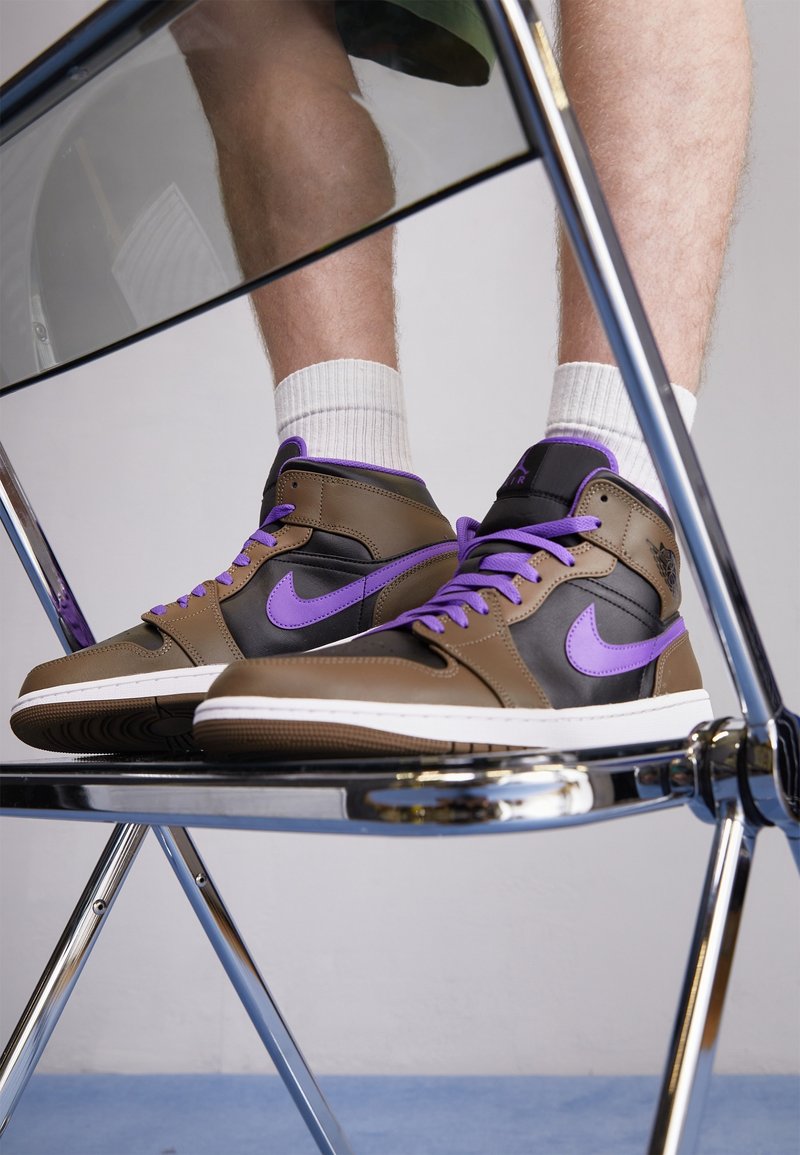 Wysokie sneakersy z brązowej skóry z czarnymi akcentami i fioletowymi detalami. Posiadają fioletowe sznurówki i białą podeszwę, opierając się na metalowym krześle.