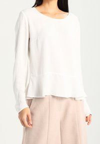 Maglietta bianca a maniche lunghe con scollatura rotonda, orlo a strati, tessuto testurizzato e silhouette morbida, abbinata a pantaloni chiari e larghi.