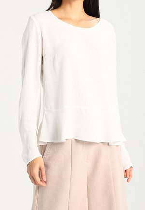 Maglietta bianca a maniche lunghe con scollatura rotonda, orlo a strati, tessuto testurizzato e silhouette morbida, abbinata a pantaloni chiari e larghi.