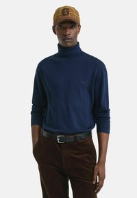 Marineblauwe coltrui van zachte stof, gecombineerd met bruine corduroy broek en een geruite pet met een groot 'G'-logo. Zwarte riem met zilveren gesp.
