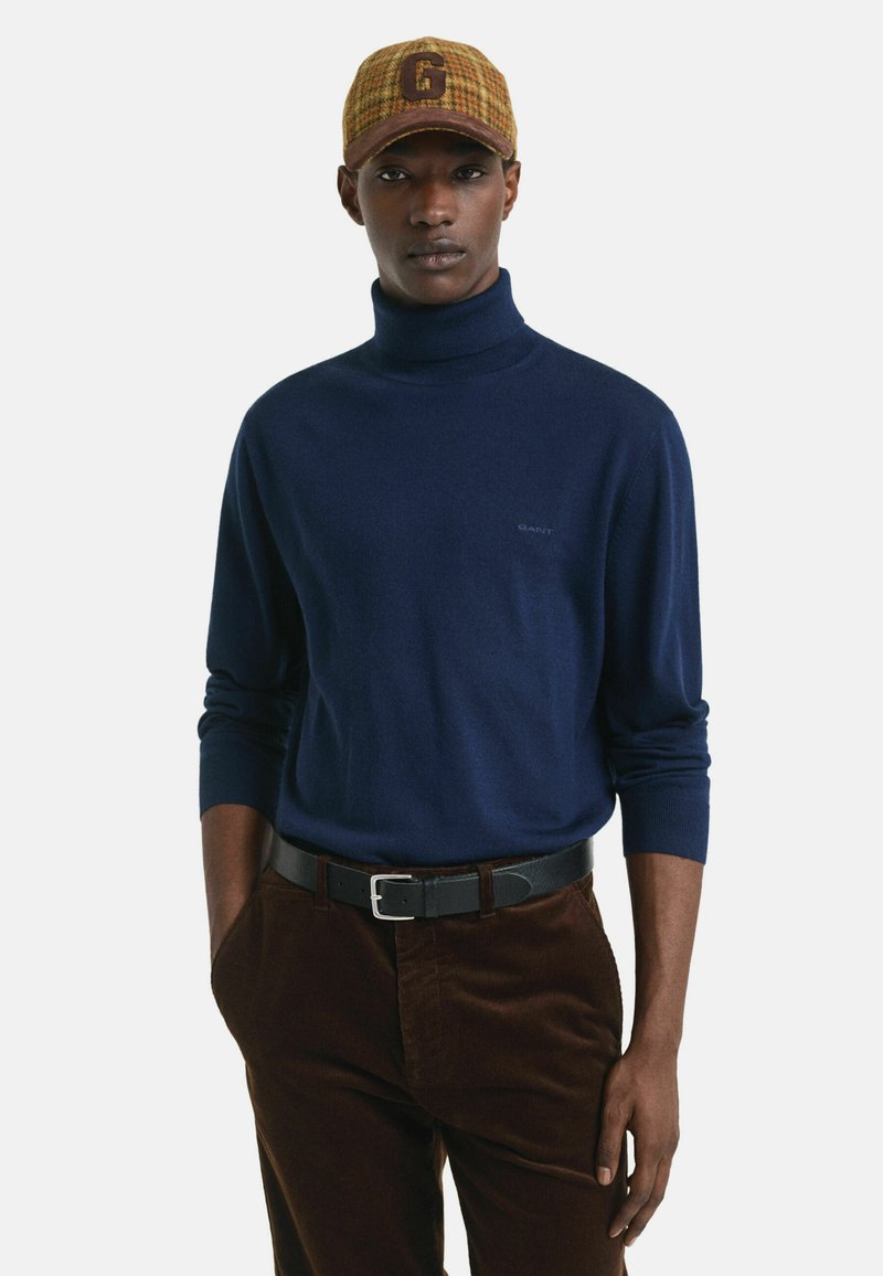 Maglione a collo alto blu navy in soffice tessuto, abbinato a pantaloni in velluto a coste marroni e un berretto a scacchi con un grande logo 'G'. Cintura nera con fibbia argentata.