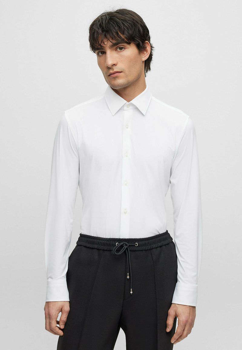 Camisa blanca de manga larga con cuello puntiagudo, hecha de tela suave, con botones al frente y puños, combinada con pantalones negros con cordón.
