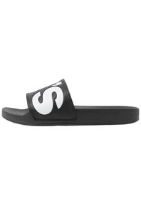 Sandale noire à glissière avec une semelle en caoutchouc texturé. Présente une large sangle avec un logo blanc proéminent sur la surface supérieure.