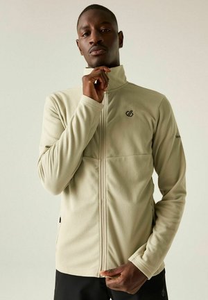 Man zipping up beige textured jacket with high collar, standing against plain light background.