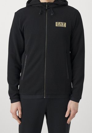 Mann trägt eine schwarze, strukturierte Zip-up-Hoodie mit Kapuze, EA7 Emporio Armani-Logo auf der Brust und seitlichen Reißverschlusstaschen.