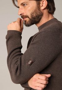 Pull en rib à côtes brun avec un col montant et un patch en cuir sur la manche. Texture douce et manches longues bien ajustées.