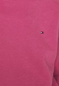 Roze katoenen trui met een geribbelde textuur en een klein geborduurd logo in rood, wit en blauw op de borst.