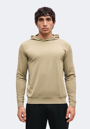 Ung mand med kort krøllet hår iført en beige hættetrøje med lange ærmer og sorte bukser, vendt mod kameraet foran en ensfarvet baggrund.