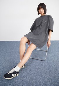 Nuori henkilö, jolla on lyhyet hiukset, istuu metallituolilla, päällään ylisuuria tummanharmaita t-paitamekkoja ja mustavalkoisia lenkkareita sinisellä matolla.