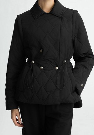 Femme portant une veste matelassée noire avec un col, des boutons argentés et des cordons réglables à la taille, debout avec une main dans la poche.