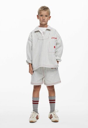 Cămașă supradimensionată în dungi albe și albastre, cu accente roșii; pantaloni scurți asortați; șosete gri cu dungi roșii; și adidași deschiși la culoare cu detalii roșii.