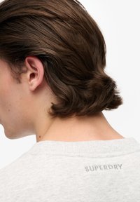 Gray sweatshirt with a textured fabric; features a ribbed neckline and silver embroidered "SUPERDRY" logo at the back near the collar.