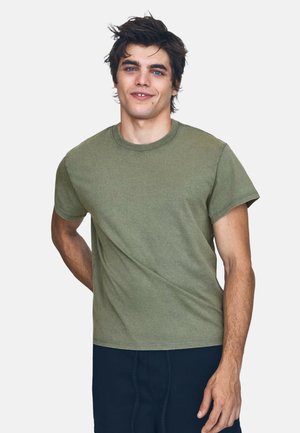 Jeune homme aux cheveux sombres en bataille, portant un t-shirt vert olive à manches courtes et un pantalon sombre, souriant avec une main dans le dos.