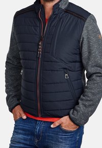 Muž oblečený v námořnické prošívané vestě s kapsami na zip přes šedou košili s dlouhým rukávem a modré džíny, s rukama v kapsách.