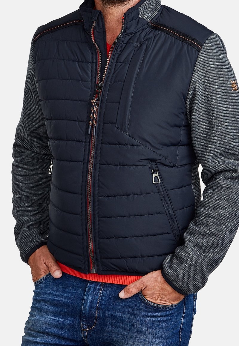 Muž oblečený v námořnické prošívané vestě s kapsami na zip přes šedou košili s dlouhým rukávem a modré džíny, s rukama v kapsách.