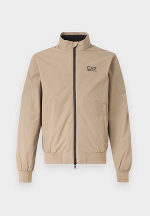 Beżowa kurtka na zamek z kołnierzem stojącym, elastycznymi mankietami, bocznymi kieszeniami oraz czarnym logo EA7 Emporio Armani na lewej piersi.