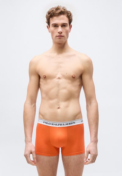 Νεαρός άνδρας με σγουρά μαλλιά στέκεται με το πρόσωπο προς τα εμπρός, φορώντας φωτεινά πορτοκαλί μπόξερ με λευκή μέση "Polo Ralph Lauren".
