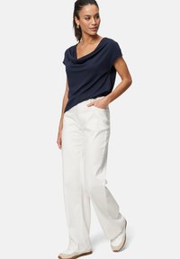 Navy cowl neck top paired with white wide-leg trousers and light-colored shoes. Fabric appears smooth; design features relaxed fit and pockets.
