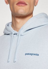 Šviesiai mėlynas džemperis su gobtuvu, pagamintas iš minkštos medžiagos, su raišteliu, įrišta apykakle ir mažuoju tamsiai mėlynu "Patagonia" logotipu ant krūtinės.