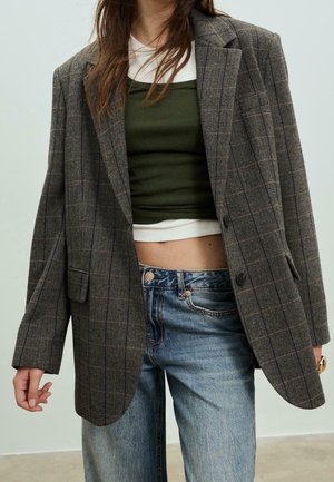 Kvinde iført oversize brun ternet blazer over grøn crop top og hvid skjorte parret med falmede blå jeans, stående mod en ensfarvet baggrund.