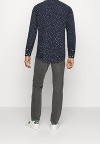 Man met een donkergepatroneerde lange mouwen shirt, grijze jeans en witte New Balance sneakers, staande tegen een eenvoudige witte achtergrond.