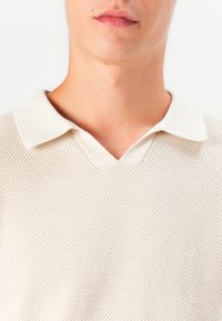 Beige strikket polo med V-hals og kontrasterende hvit krage, med et teksturert stoff med mesh-mønster.
