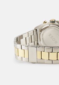 Reloj de acero inoxidable con pulsera en tonos plateado y dorado. Presenta una caja redonda, una corona texturizada y un cierre pulido.