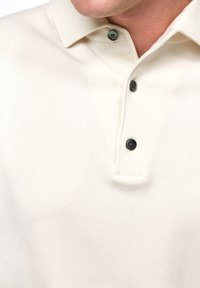 Witte polo met een zachte textuur, colkraag en drie zwarte knopen met groene accenten bij de kraag.