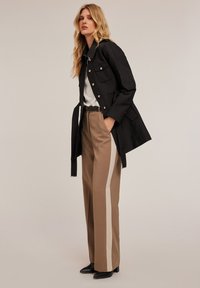 Gabardina negra con botones plateados, llevada sobre una blusa blanca y combinada con pantalones de tiro alto beige con franjas laterales color crema. Zapatos negros de punta.