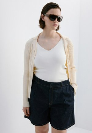 Personne portant des lunettes de soleil foncées, un cardigan crème côtelé, un haut blanc à col en V et un short taille haute foncé, debout devant un fond blanc.