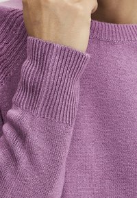 Lila Strickpullover mit geripptem Kragen und Bündchen, glatter Textur und figurbetontem Design. Nahaufnahme des Ärmel-Details.