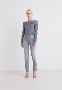 Top de punto acanalado off-the-shoulder en azul oscuro, combinado con jeans ajustados gris claro y sandalias plateadas de tiras, contra un fondo blanco.