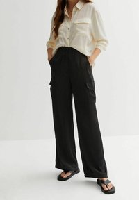 Woman wearing a cream button-up shirt tucked into high-waisted black wide-leg pants with side pockets and black sandals.