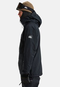 Schwarze wasserdichte Jacke mit verstellbarem Kapuzenaufbau, Reißverschlusstaschen und seitlichem Logo. Getragen mit einer braunen Strickmütze und schwarzen Handschuhen.