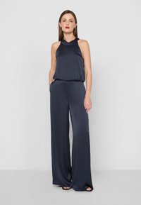 Námornícky halter overall z hladkej tkaniny pripomínajúcej satén, so širokými nohavicami, voľným strihom a bočnými vreckami pre zvýšenú funkcionalitu.