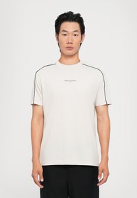 Camiseta ligera beige con cuello redondo, mangas cortas y acentos en negro en los hombros. Presenta un logo central en texto pequeño. Textura suave.