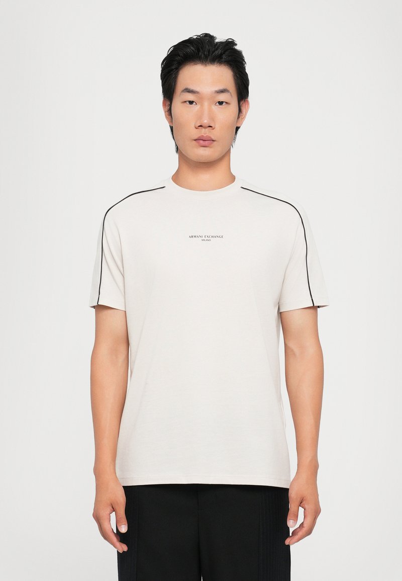 Camiseta ligera beige con cuello redondo, mangas cortas y acentos en negro en los hombros. Presenta un logo central en texto pequeño. Textura suave.