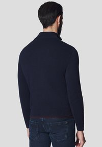 Granatowy sweter z dzianiny z wysokim kołnierzem i prążkowanymi mankietami, z wyróżniającą się fakturą. Noszony z ciemnymi dżinsami o subtelnym wzorze.