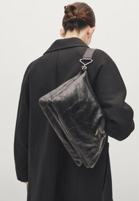 Sac en cuir noir avec une texture lisse, de forme rectangulaire et avec des finitions en métal argenté, porté sur une bandoulière. Le modèle porte un manteau noir.