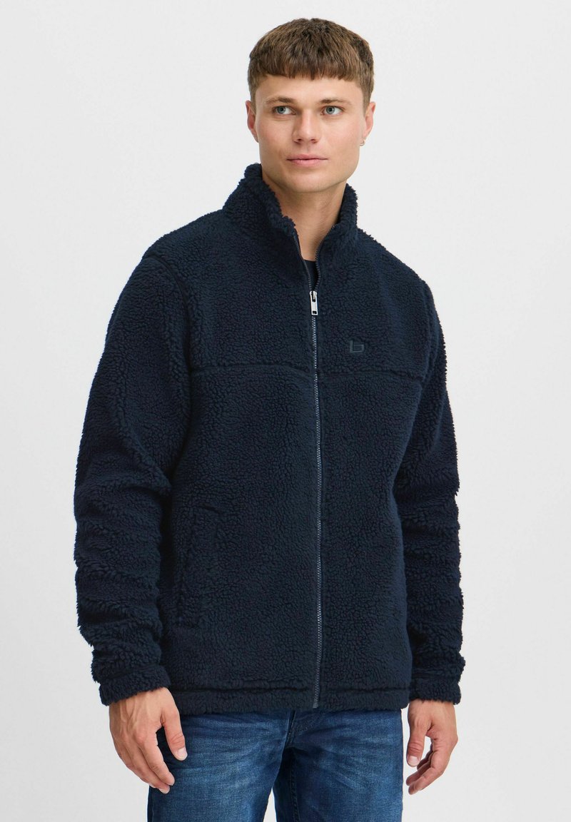 Veste en polaire navy avec un col haut, fermeture éclair intégrale, surface texturée et poches latérales. Caractéristiques d'un logo sur la poitrine et de poignets élastiques.