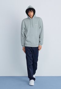 Felpa grigia con tasca frontale, stile kangaroo, cappuccio regolabile con cordino, indossata con pantaloni blu navy e scarpe bianche, dettagli di branding minimi.