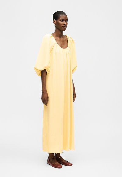 Γυναίκα όρθια που φοράει ένα μακρύ, φαρδύ φόρεμα ανοιχτό κίτρινο με φουσκωτά μανίκια μέχρι τον αγκώνα και καφέ επίπεδα παπούτσια.