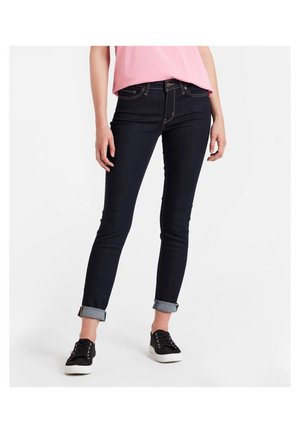 Slim Fit dunkelblaue Jeans, an den Knöcheln aufgerollt, getragen mit einem pinken Oberteil und schwarzen Low-Top-Sneakern vor einem schlichten weißen Hintergrund.