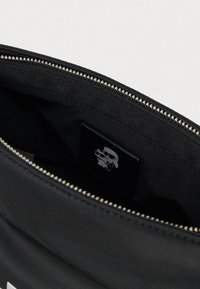 Μαύρη τσάντα υφάσματος με κλείσιμο φερμουάρ, με λεία υφή. Στο εσωτερικό φαίνεται μια μικρή γραφική ετικέτα και ελάχιστες εσωτερικές λεπτομέρειες.