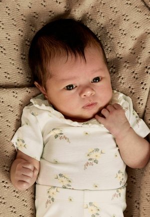 Nyfødt baby med mørkt hår som har på seg et hvitt blomsterantrekk, ligger på et teksturert beige teppe og ser rolig mot kameraet.